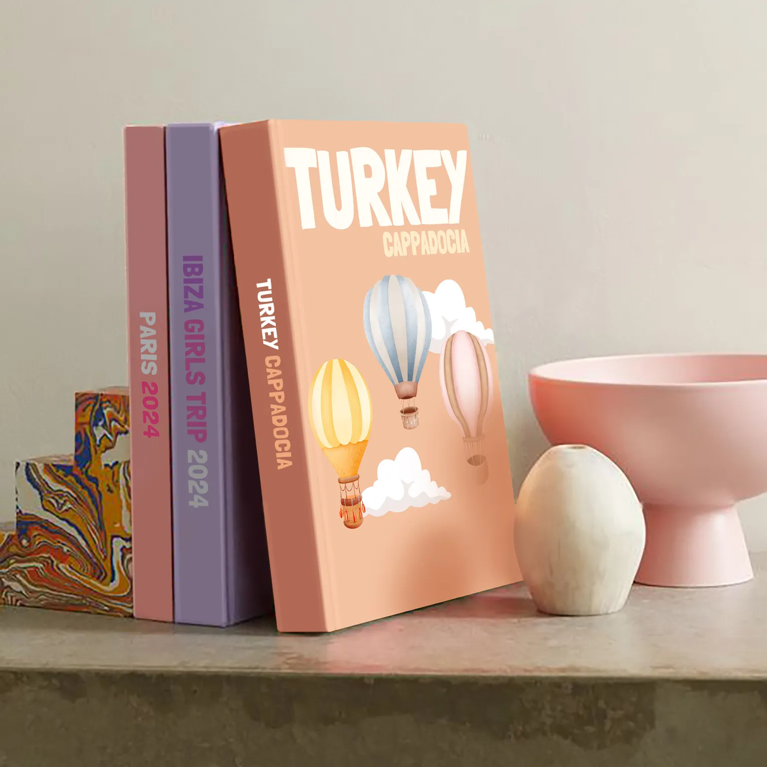 Three Kito travel books on a concrete shelf, spines reading Paris 2024 and Ibiza Girls Trip 2024, the front one a Turkey Cappadocia cover with hot-air balloons drifting across it.