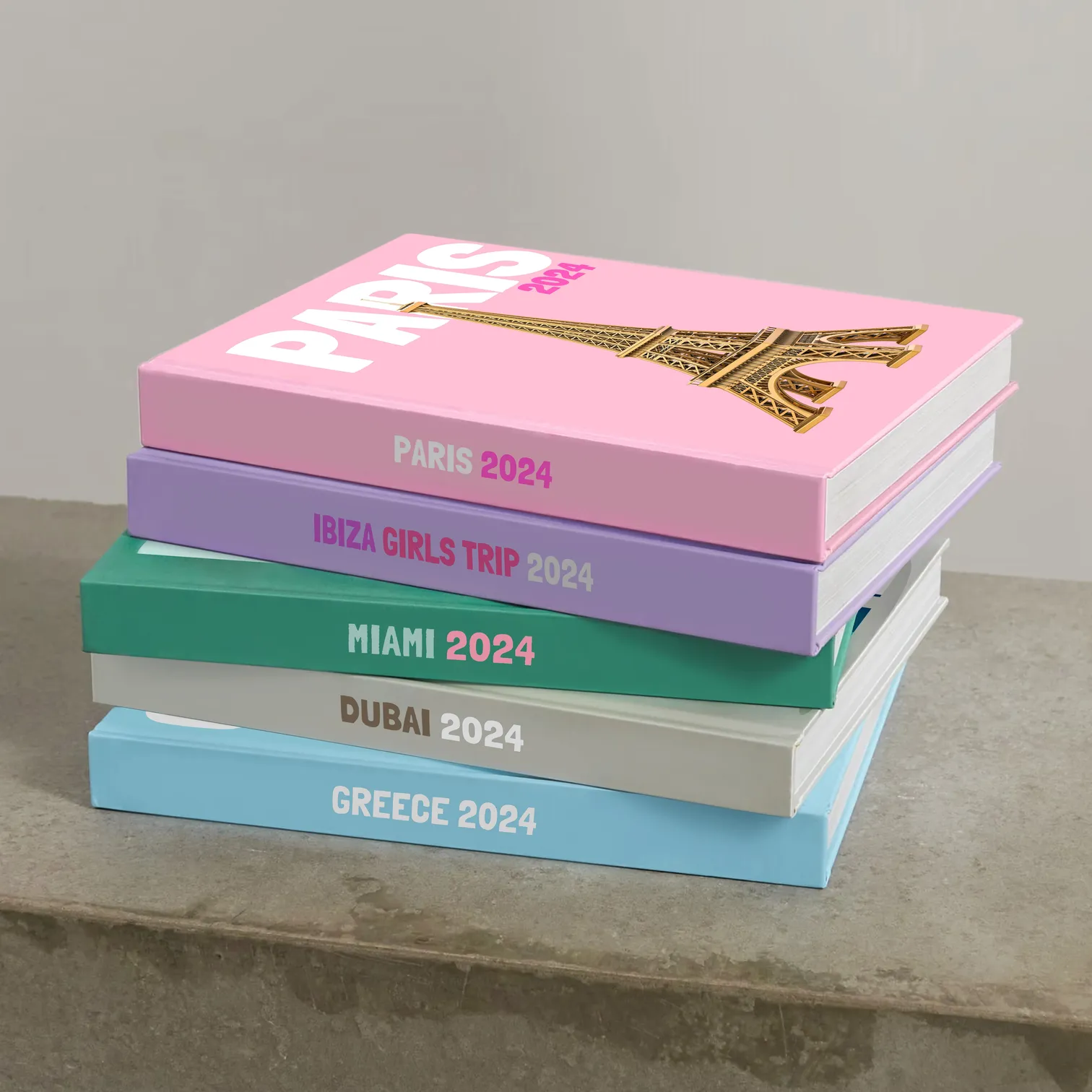 Five Kito travel books stacked on a concrete ledge, spines reading Paris, Ibiza Girls Trip, Miami, Dubai and Greece, the top one a pink Paris cover with a gold Eiffel Tower.